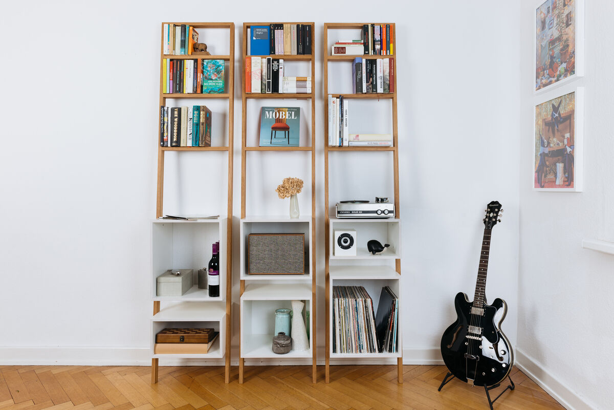 Ein modernes Leiterregal aus Holz und weißen Elementen, gefüllt mit Büchern, Dekorobjekten und elektronischen Geräten, steht an einer weißen Wand. Ein schwarzes Musikinstrument lehnt an der rechten Seite.