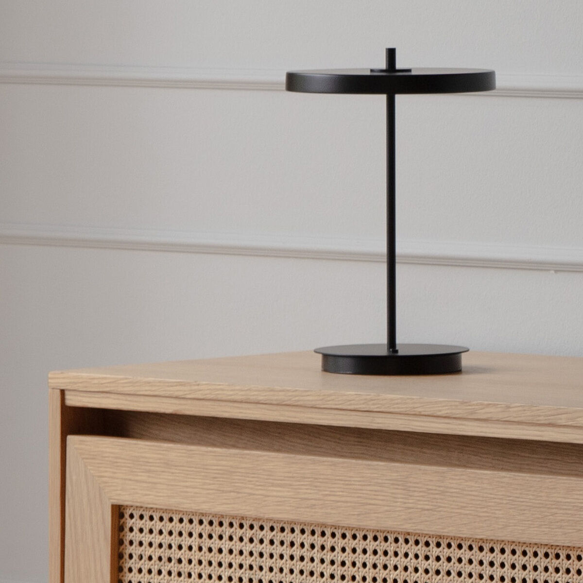Moderne, kabellose schwarze Tischlampe Asteria Move steht auf einem hellen Holzmöbel mit geflochtenem Einsatz vor einer schlichten, grau-weißen Wand.