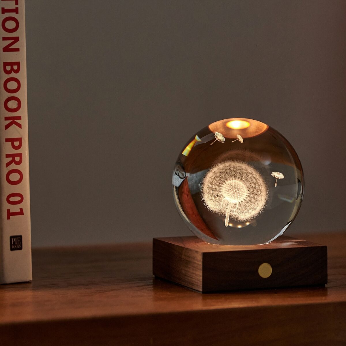 Eine elegante Lampe »Amber Light« von Gingko Design steht auf einem Sideboard. Die runde Kristallglaskugel mit filigranem, leuchtendem Pusteblumenmotiv ruht auf einem edlen Sockel aus dunklem Walnussholz mit goldfarbenem Bedienknopf. Im Hintergrund sind Bücher, ein orangefarbenes Kissen und eine Brille sichtbar.