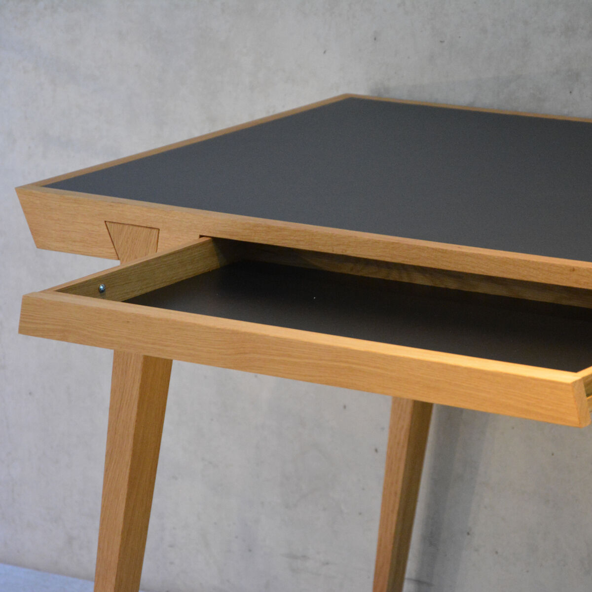 Ein moderner Schreibtisch aus Holz mit einer schwarzen Oberfläche und einer Schublade. Der Tisch hat vier Beine und ist schlicht gestaltet. Der Hintergrund ist neutral und graufarben.