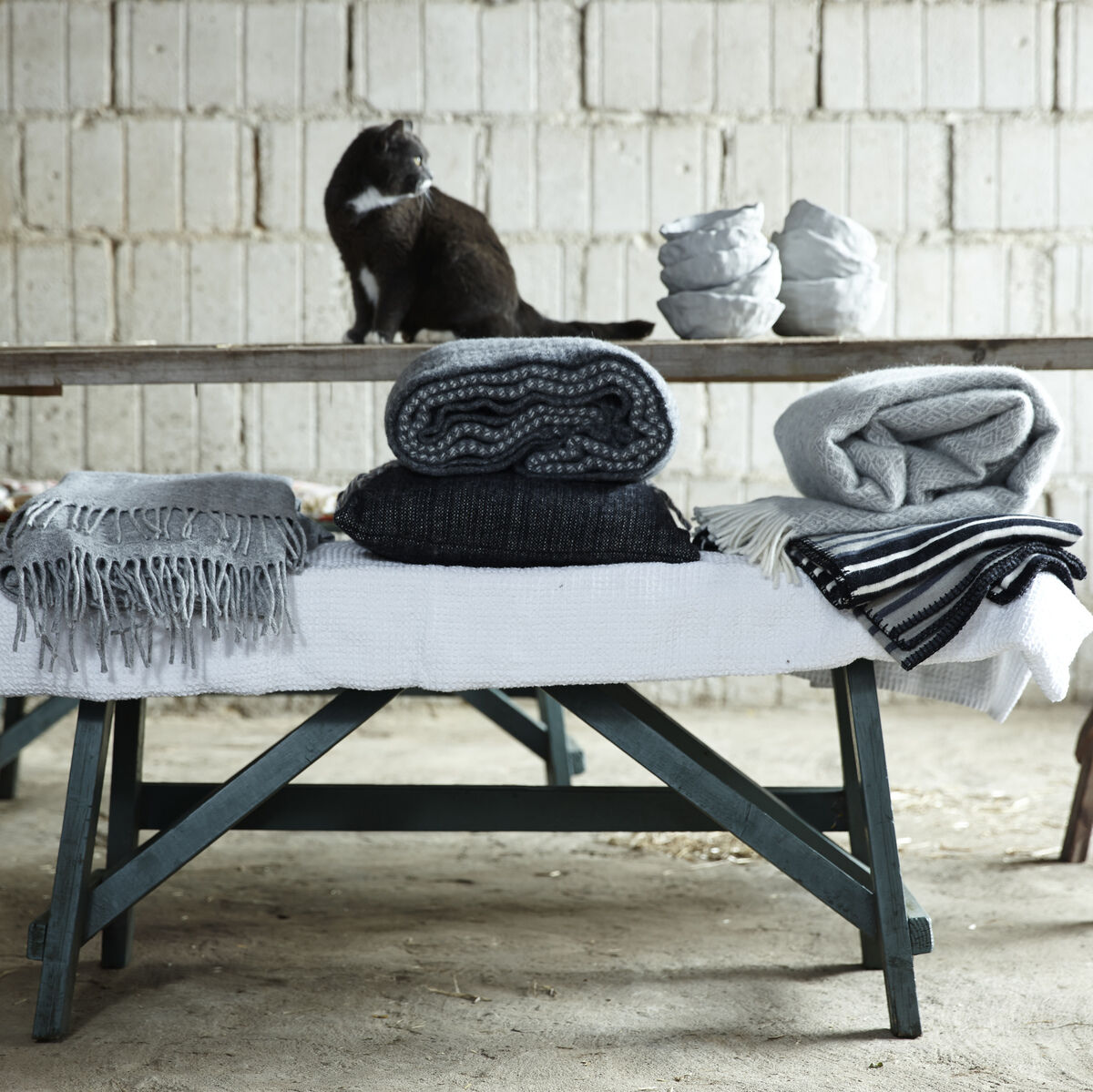 Eine gemütliche, skandinavisch inspirierte Szene mit gefalteten Decken in Grau- und Weißtönen auf einem rustikalen Holztisch. Im Hintergrund eine Katze und stapelbare Schalen vor einer Steinwand.