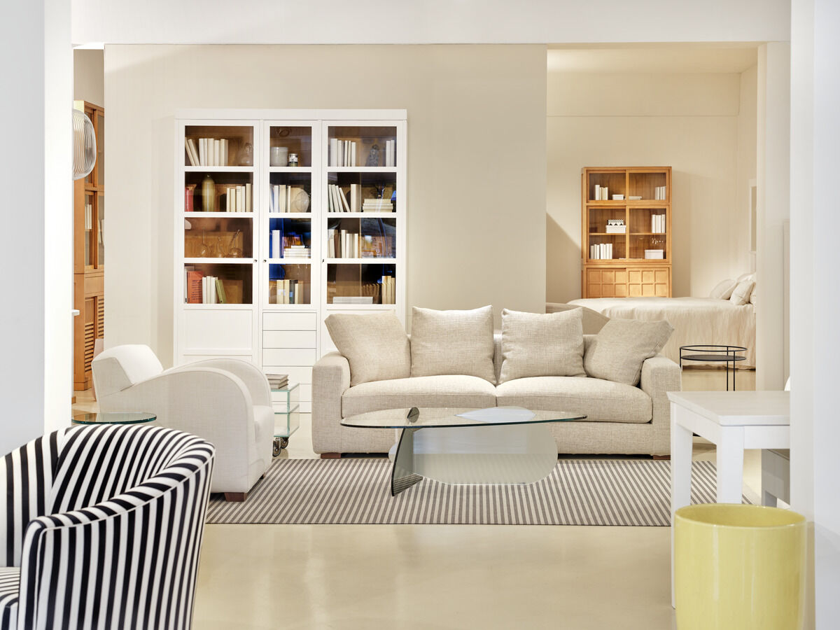 Helles und modernes Wohnzimmer mit einem weißen Sofa. Im Hintergrund befinden sich eine weiße Vitrine und ein Bücherregal mit Büchern. Vor dem Sofa liegt ein grauer Teppich, und ein Couchtisch aus Glas steht darauf. Ein gemütlich eingerichteter Raum mit klaren Linien und eleganter Dekoration.