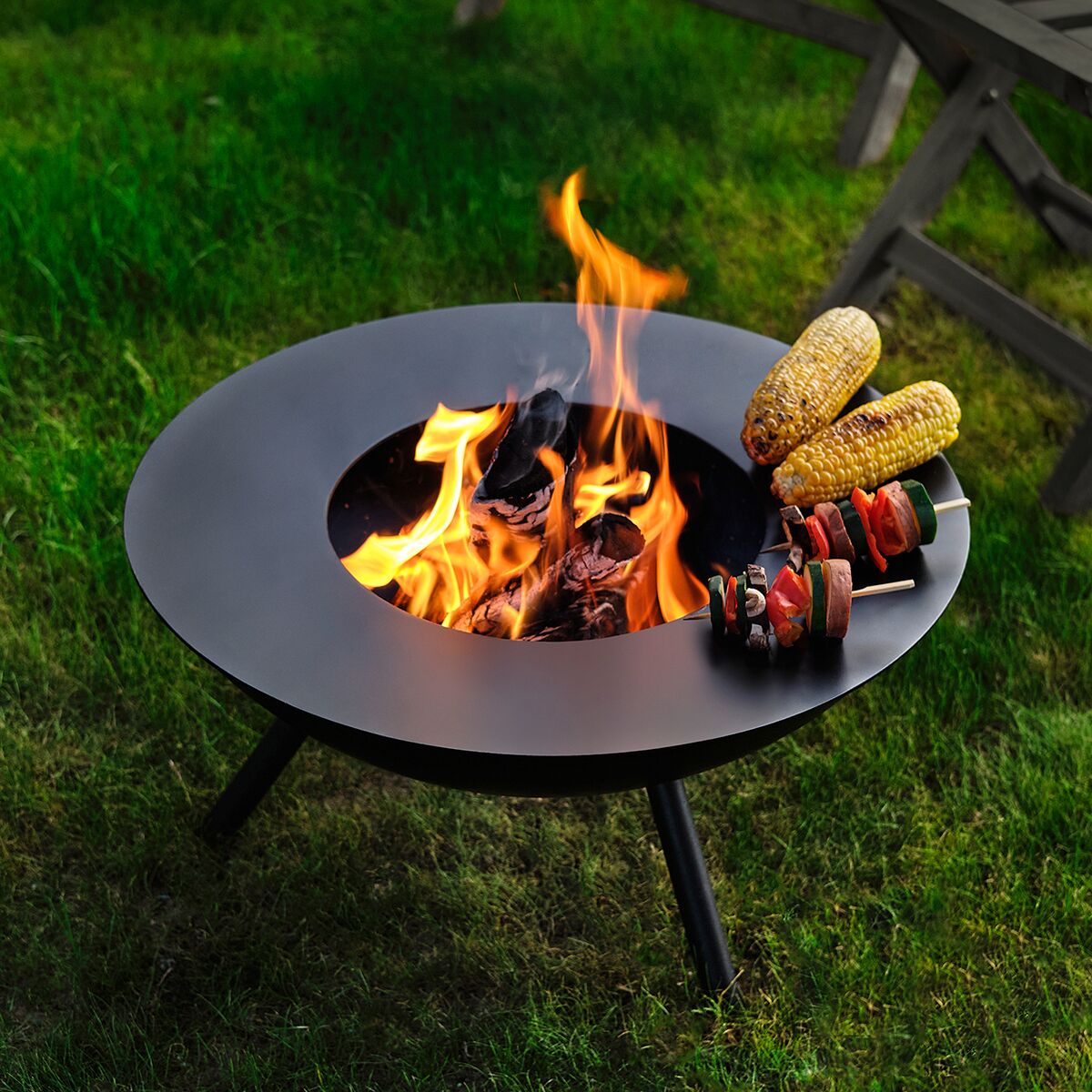 Im Mittelpunkt steht eine runde, mattschwarze Grillplatte »Flames« von Philippi Lager auf einer Feuerschale im Garten. Über offener Flamme liegen zwei Maiskolben und zwei Spieße mit Gemüse und Würstchen auf der Grillplatte. Grün leuchtender Rasen bildet den Hintergrund; rechts ein Holztisch.