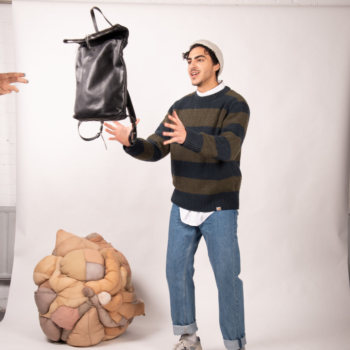 Ein junger Mann, der eine Mütze, einen gestreiften Pullover, ein Hemd und Jeans trägt, fängt lächelnd einen schwarzen Rucksack namens »Nabuku«. Im Hintergrund befindet sich eine farbige Struktur auf einem weißen Boden vor einer weißen Wand.