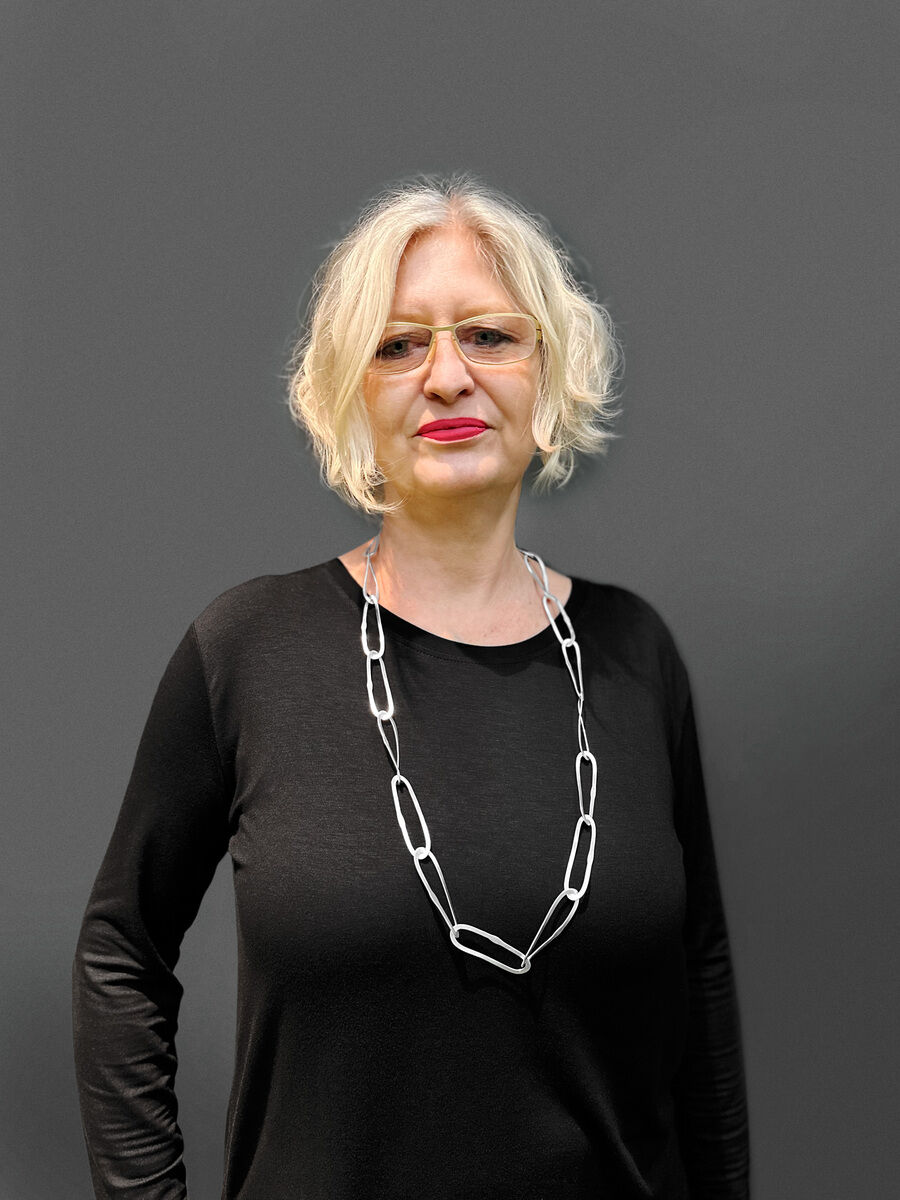 Eine Frau trägt eine Silberkette mit wellenförmigen Anhängern. Sie hat blondes, schulterlanges Haar, eine Brille und trägt ein schwarzes Oberteil.