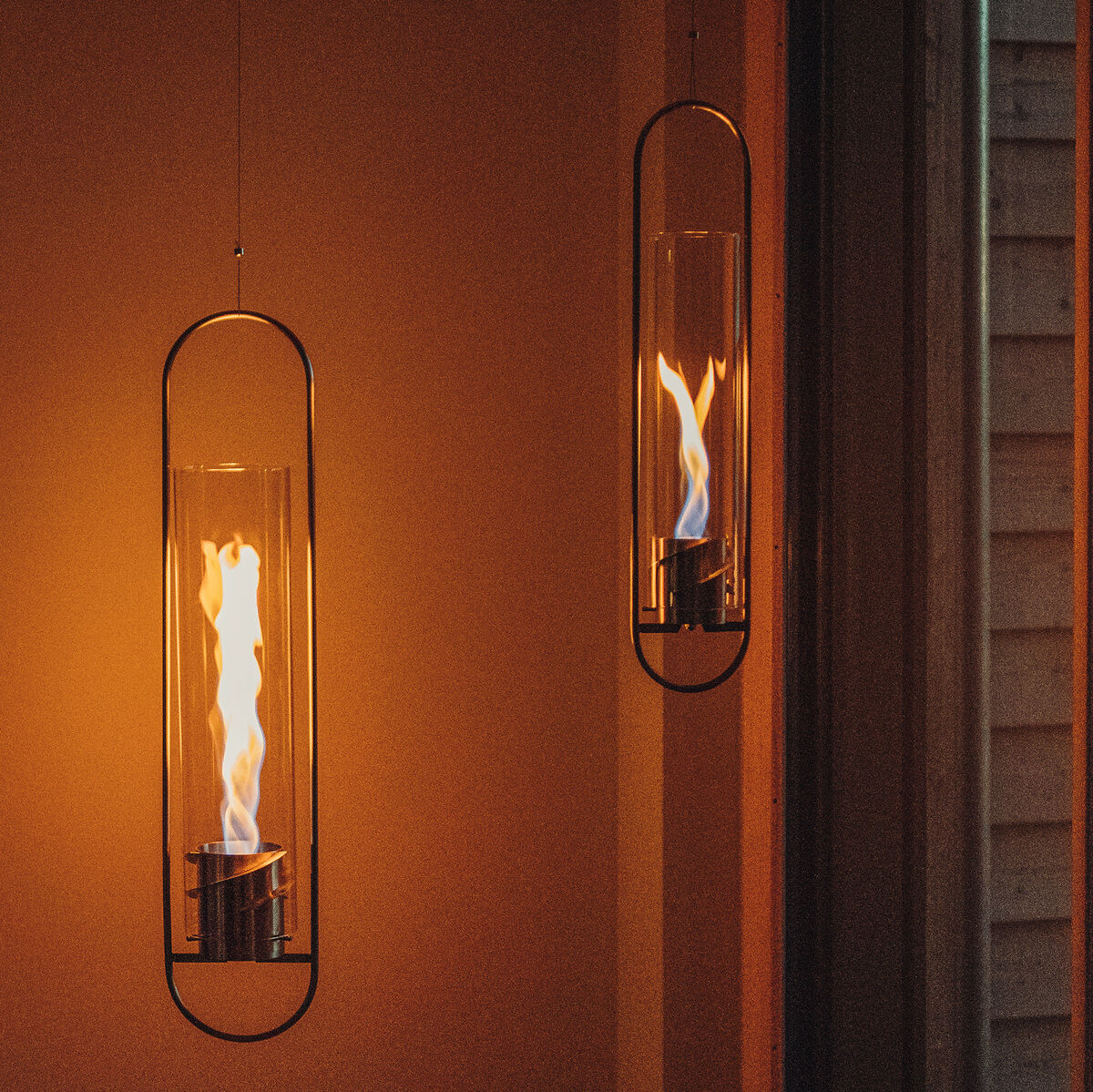 Zwei moderne Hängelampen aus Glas, in denen Flammen leuchten, hängen in einem gemütlich beleuchteten Raum. Die Lampen vermitteln eine warme und entspannte Atmosphäre.
