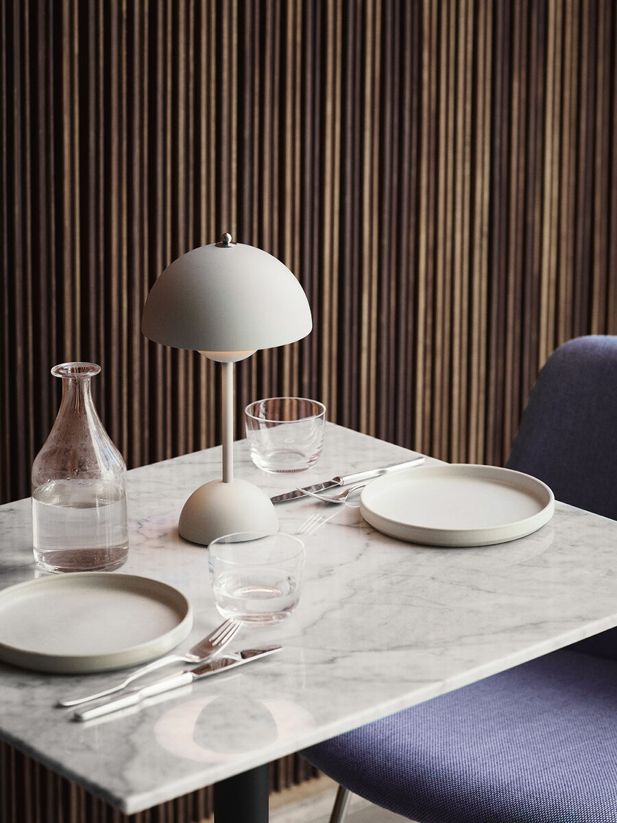 Eine moderne Tischleuchte mit Akku, die auf einem Esstisch mit Marmordekor steht. Die Leuchte hat einen runden weißen Schirm und ein minimalistisches Design. Im Hintergrund befinden sich eine graue Wand und ein gepolsterter Stuhl. Auf dem Tisch stehen zwei Teller, zwei Gläser, eine Glasflasche und Besteck.