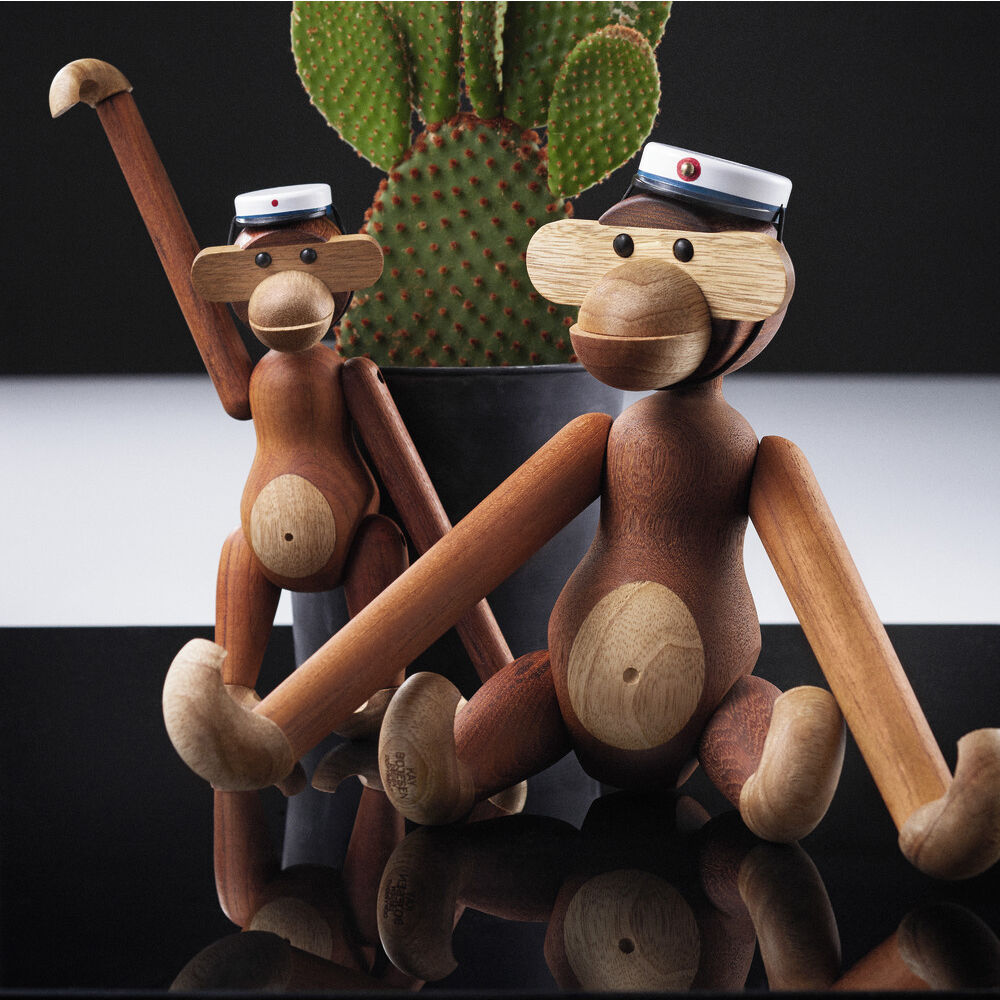 Zwei Holzfiguren in Form von Affen der Marke Kay Bojesen sitzen und lehnen aneinander vor einem grünen Kaktus. Die Affen sind aus Teak- und Limbaholz gefertigt und tragen weiße Graduierungsmützen. Glänzende schwarze Fläche spiegelt die Szene.