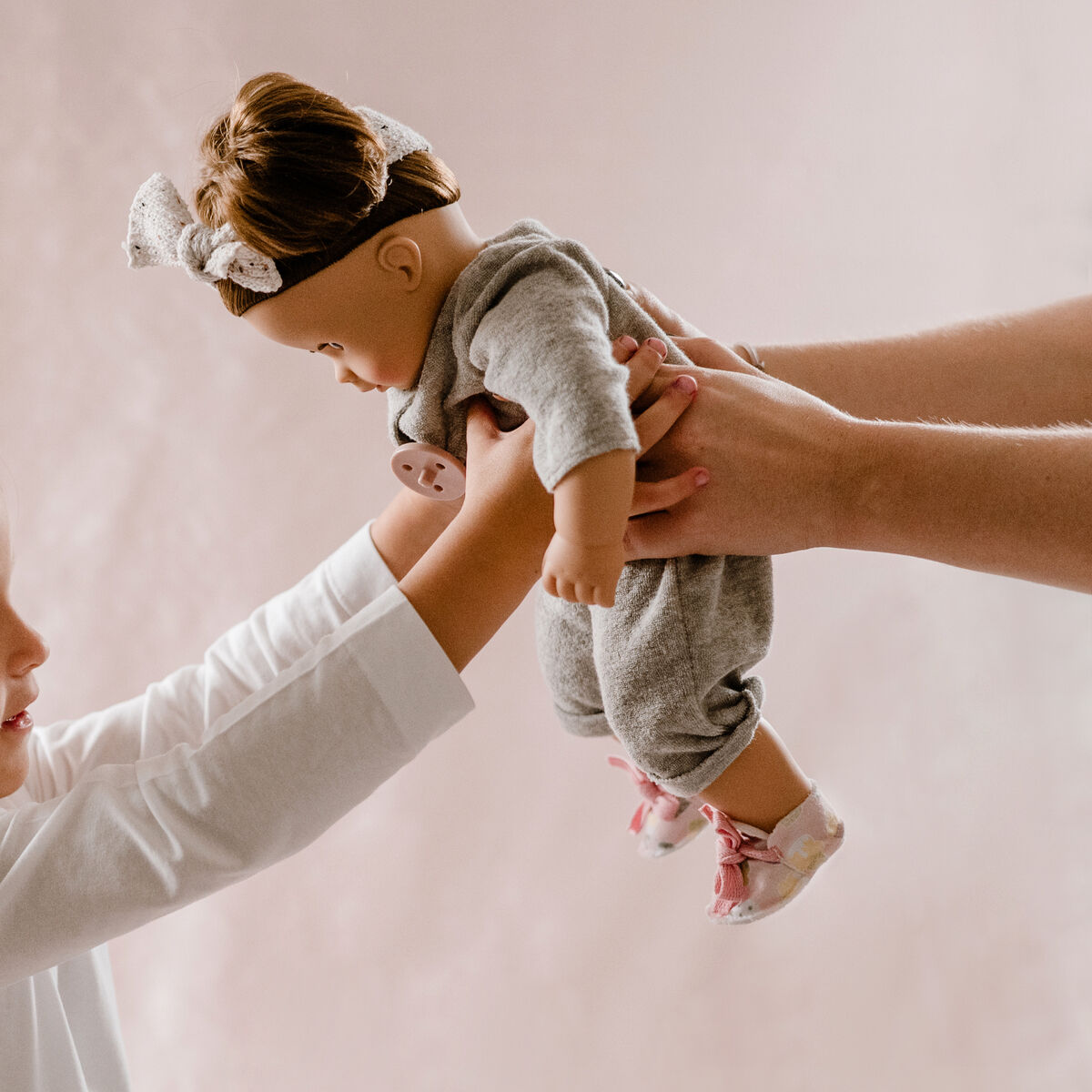 Ein junges Mädchen in weißem Oberteil hält eine Stoffpuppe namens »Maxy Muffin Popsicle«, während ein Erwachsener die Puppe übergibt. Die Szene zeigt die Freude des Kindes beim Erhalten des Spielzeugs, im Hintergrund ein weicher, heller Hintergrund.