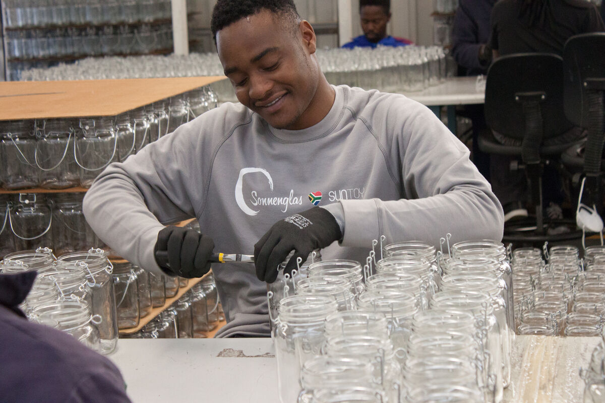 Ein Mann sitzt und montiert ein Sonnenglas, umgeben von vielen ähnlichen Gläsern in einem industriellen Arbeitsumfeld.