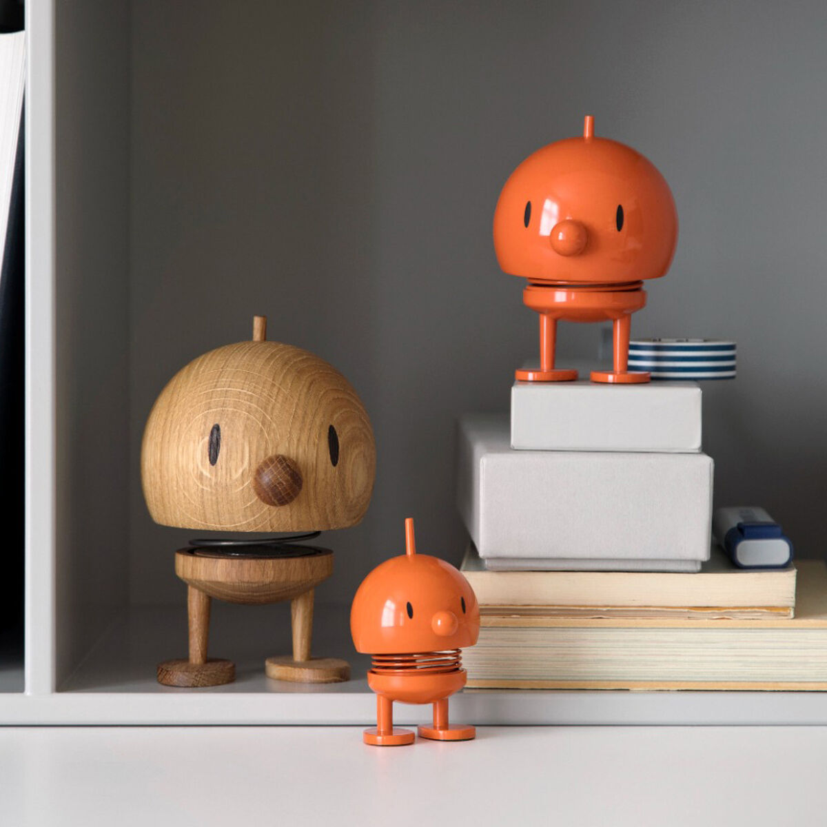 Drei Hoptimist Bumble-Figuren aus Eiche und orangefarbenem Kunststoff stehen in einem minimalistischen Regal, umgeben von Büchern und anderen kleinen Dekorationsobjekten.