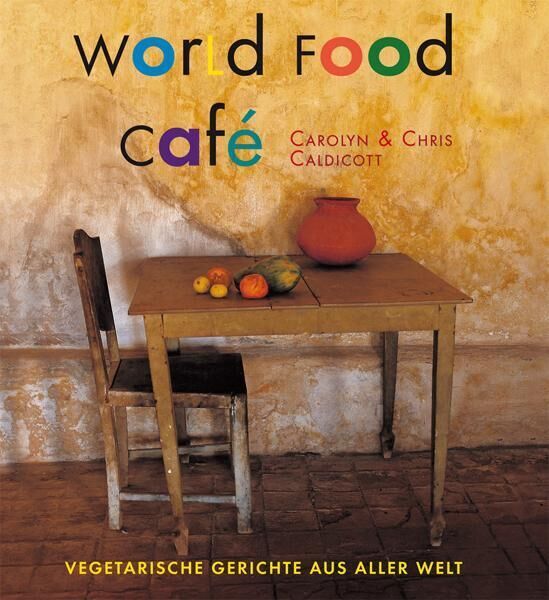 Das Cover des Buches 'World Food Café' von Carolyn und Chris Caldicott zeigt einen rustikalen Holztisch mit Obst und einer Tonvase. Ein einfacher Stuhl steht neben dem Tisch, der vor einer beigen, leicht abgenutzten Wand steht. Der Titel ist in bunten Buchstaben und der Untertitel lautet 'Vegetarische Gerichte aus aller Welt'.