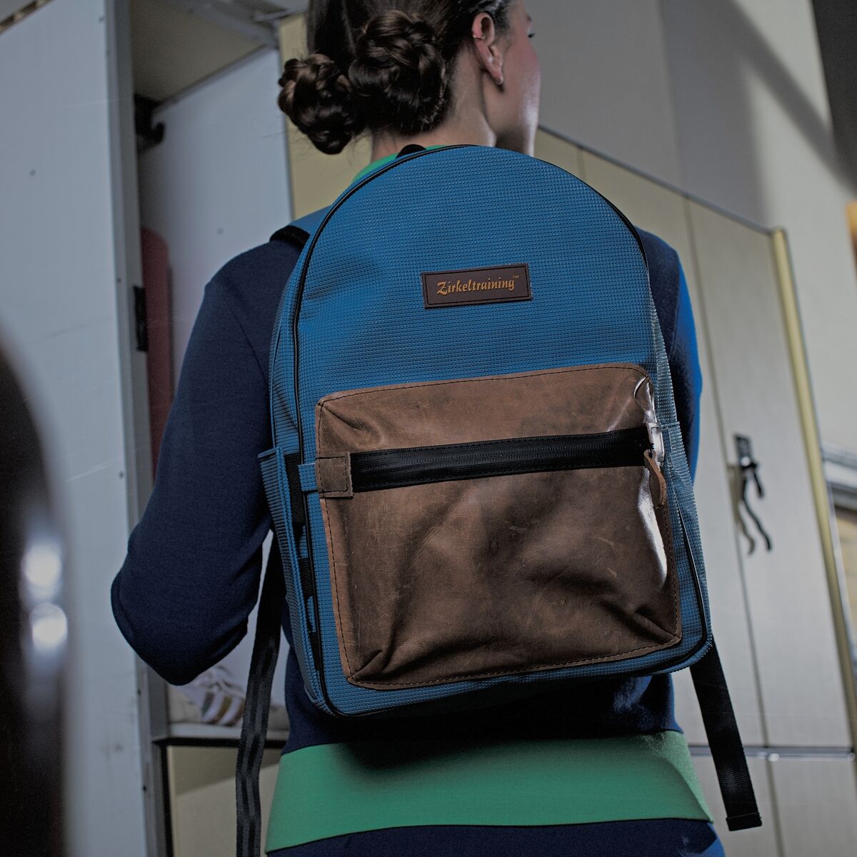 Junge Frau trägt einen blauen Rucksack mit braunen Akzenten. Der Rucksack hat einen markanten Frontreißverschluss und wird in einer sportlichen Umgebung getragen.