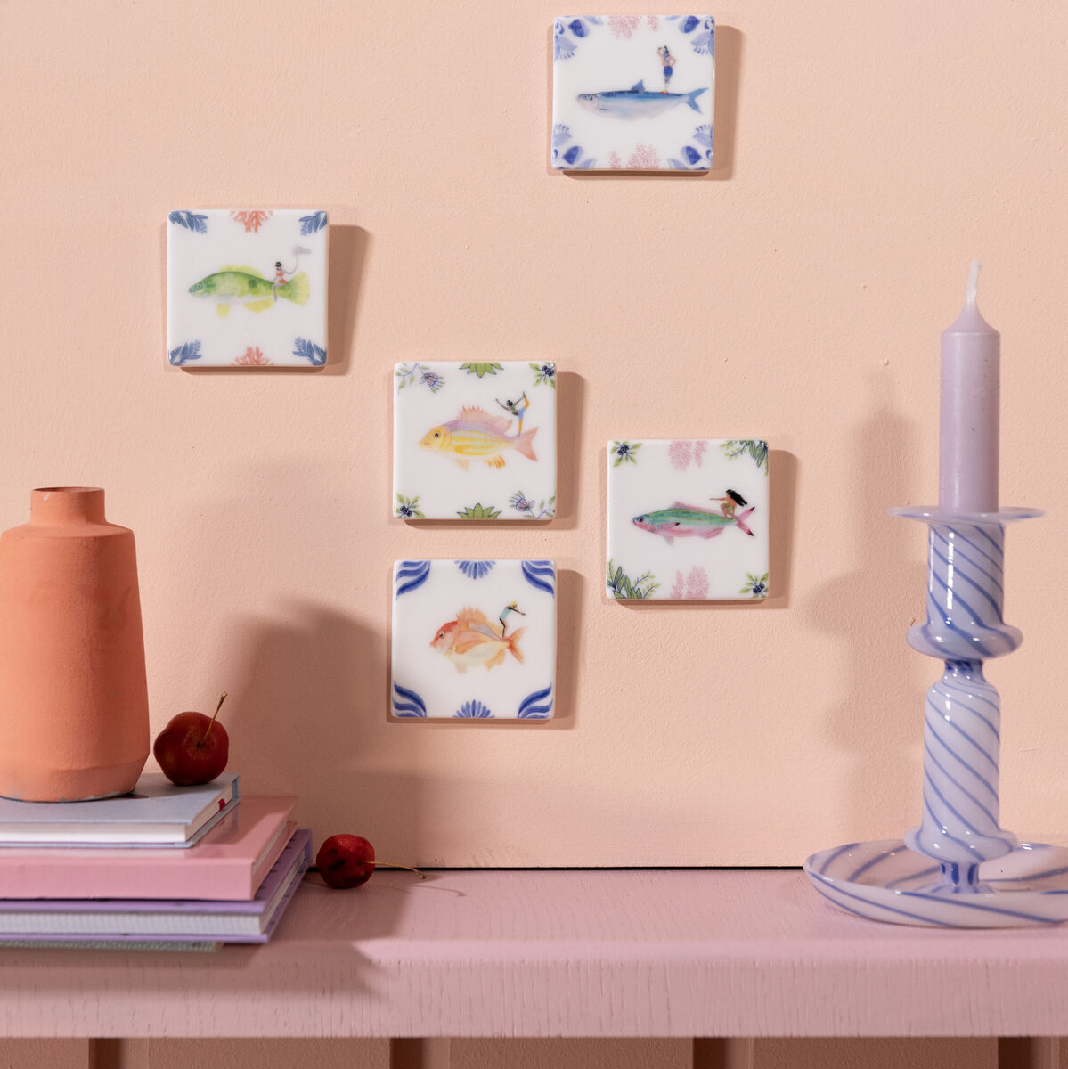 Fünf Keramikfliesen mit verschiedenen Tiermotiven sind an einer rosa Wand angebracht. Eine weiße Kerze in einem gestreiften Kerzenständer und eine orangefarbene Vase mit Büchern und einer Kirsche sind unten auf einem Regal platziert. Die Fliesen sind in Pastellfarben gehalten und zeigen springende Tiere wie Fische, Schildkröten und Frösche.