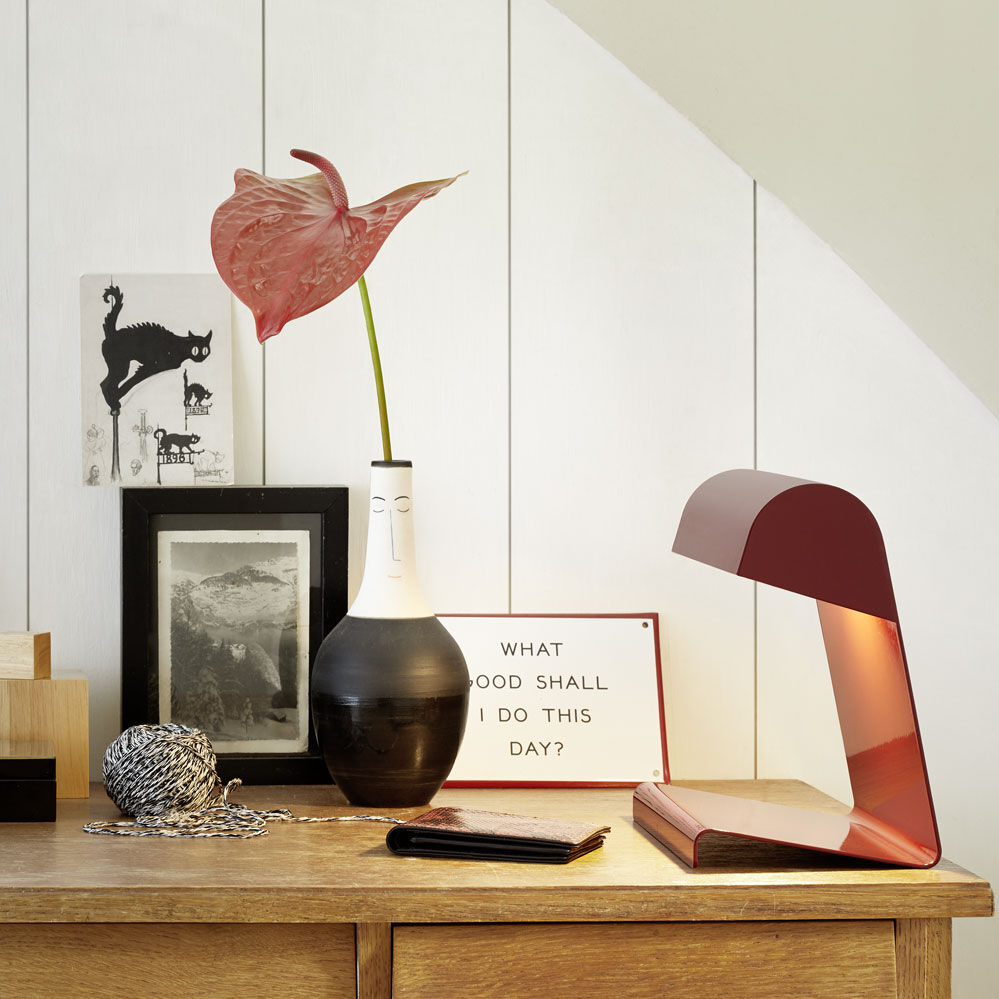Ein modernes Sideboard aus Holz steht vor einer weiß vertäfelten Wand. Darauf steht rechts die Tischlampe »Lampe de Bureau« von Vitra in tiefschwarzer und japanisch roter Ausführung, daneben eine schwarz-weiße Vase mit einer rosa Blume, dekorative Wandbilder und Büromaterial.
