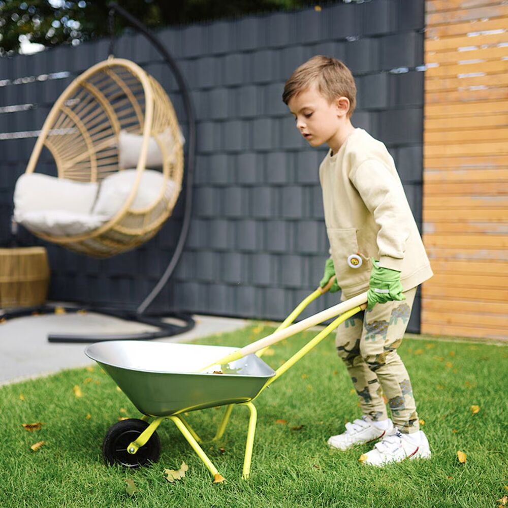 Ein Kind schiebt eine gelbgrüne Kinder-Schubkarre mit Gartenwerkzeug auf einer Rasenfläche. Das Kind trägt Gartenhandschuhe, einen beigen Pullover und Camouflage-Hose. Im Hintergrund sind ein Hängesessel und ein moderner Holzzaun zu sehen.