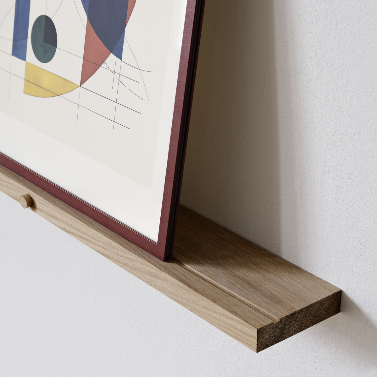 Ein Galerie Regal aus Eichenholz an einer weißen Wand. Auf dem Regal steht ein bunter gerahmter Kunstdruck.
