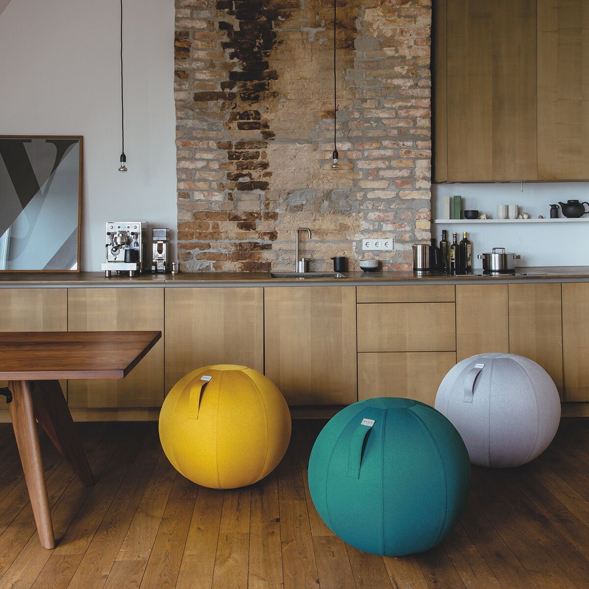 Drei Sitzbälle der Marke »Vluv Leiv« in den Farben Gelb, Grün und Grau befinden sich in einem modernen, gemütlichen Raum mit Holzboden und Backsteinwand. Ein hölzerner Tisch und eine Küche sind im Hintergrund zu sehen.