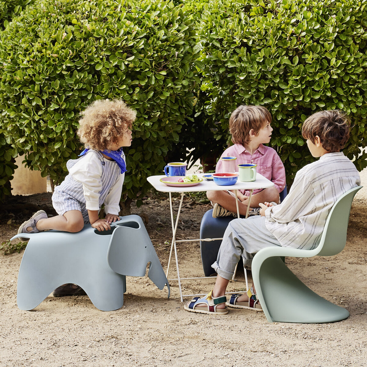 Mehrere Kinder spielen und sitzen auf modernen, farbenfrohen Kindermöbeln, darunter der graue Eames Elephant Hocker, im Garten. Erwachsene sind auch anwesend im Hintergrund. Die Szene spielt draußen an einem sonnigen Tag und ist von grüner Vegetation umgeben.