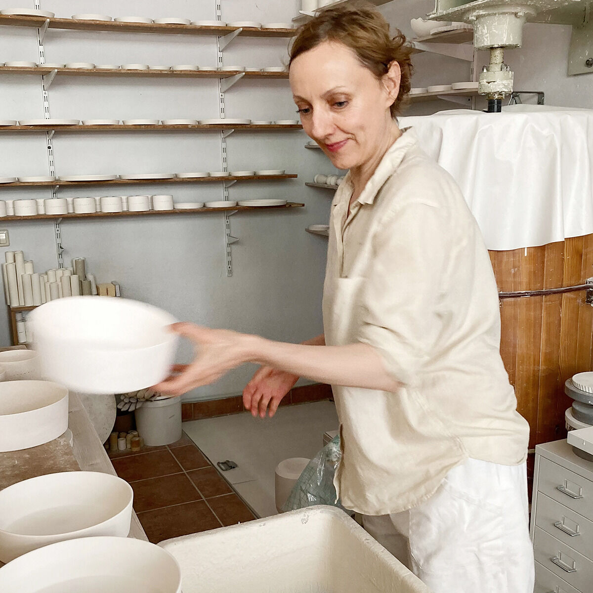 Eine Person in einem Atelier hält einen Kerzenhalter aus Porzellan. Im Hintergrund sind weitere Keramikgegenstände sichtbar.