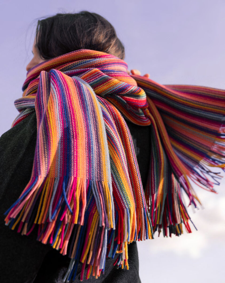 Eine Person trägt einen großen, auffällig gestreiften Merinoschal in Rot, Blau, Gelb, Orange und weiteren bunten Farben. Der Schal hat lange, bunte Fransen und wirkt weich und wärmend. Die Person steht im Freien, vor einem hellen Himmel.