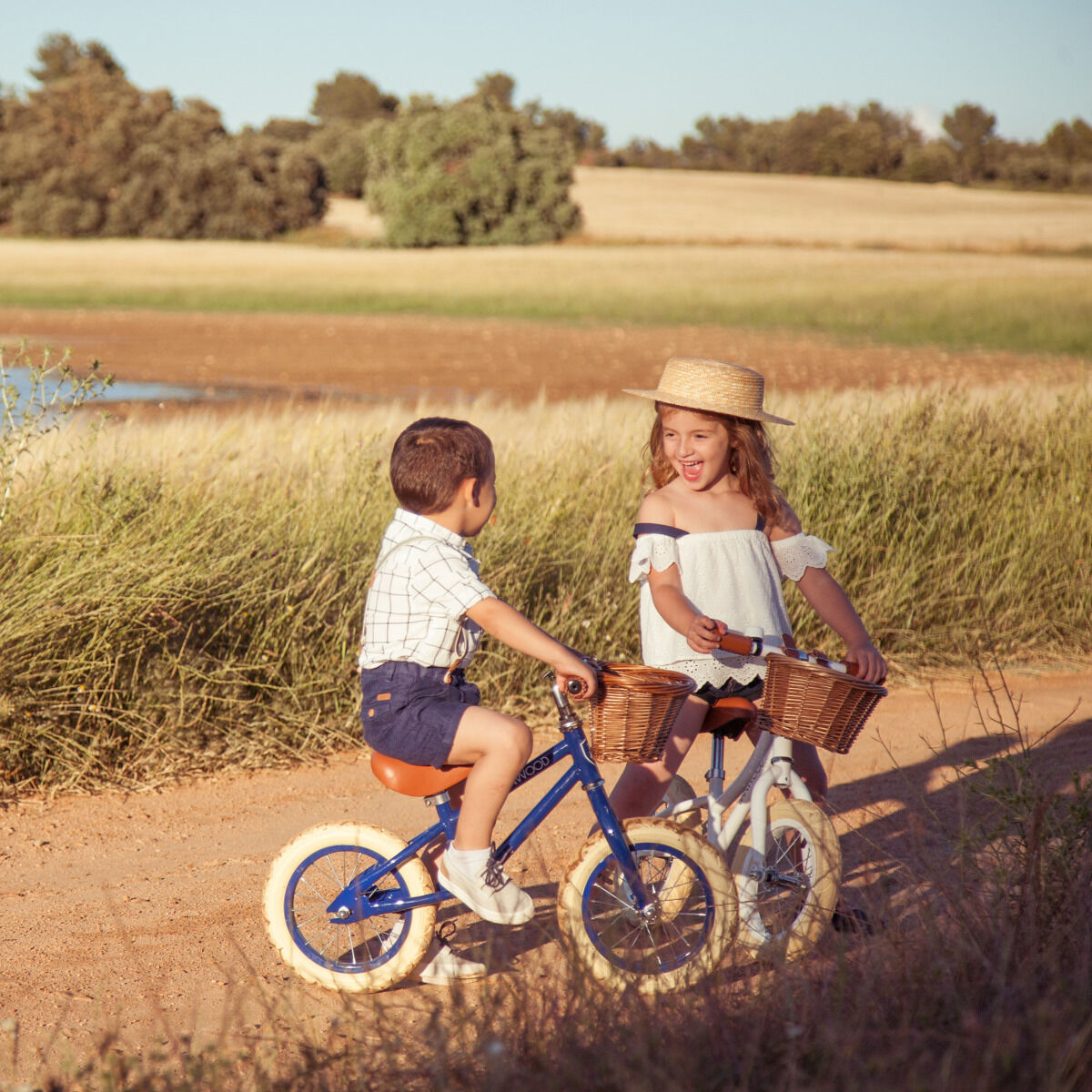 Ein Junge und ein Mädchen fahren auf einem Weg in der Natur auf Retro-Laufrädern mit kleinen Holzkörbchen vorne. Der Junge trägt ein blau-weiß gestreiftes T-Shirt und blaue Shorts, das Mädchen ein weißes Oberteil und einen Strohhut. Im Hintergrund sind grüne Wiesen und Bäume zu sehen, unter einem klaren Sommerhimmel.