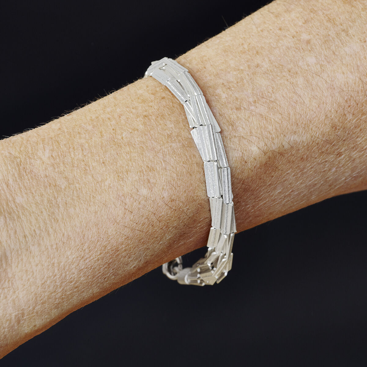 Nahaufnahme eines geprägten Silberarmbands am Handgelenk vor schwarzem Hintergrund. Das stilvolle Schmuckstück zeigt ein modernes, zeitloses Design und eignet sich für festliche Anlässe.