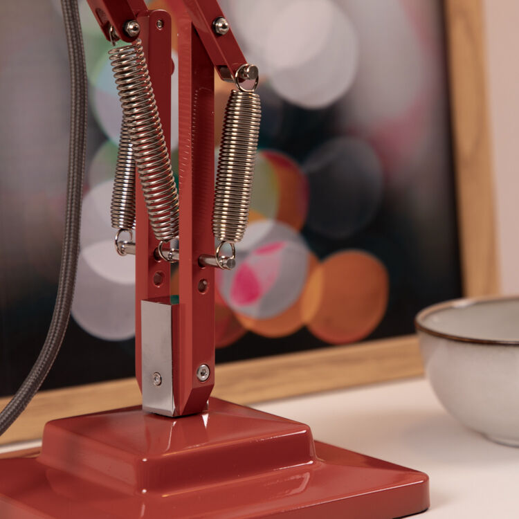 Nahaufnahme einer roten Tischleuchte der ZEIT-Sonderedition »Anglepoise Original 1227», die auf einem Tisch neben einer Schüssel steht. Im Hintergrund ist ein gerahmtes Bild mit unscharfen, bunten Lichtern zu sehen.