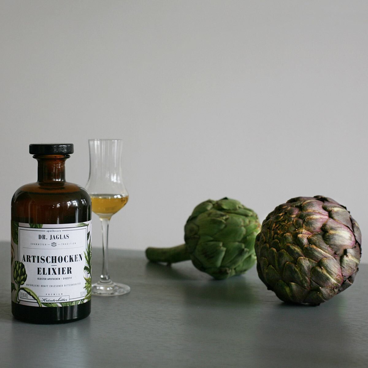 Eine braune Flasche »Artischocken-Elixier (Kräuterbitter)« von Dr. Jaglas steht auf einem Tisch neben zwei Artischocken und einem halb gefüllten Glas.