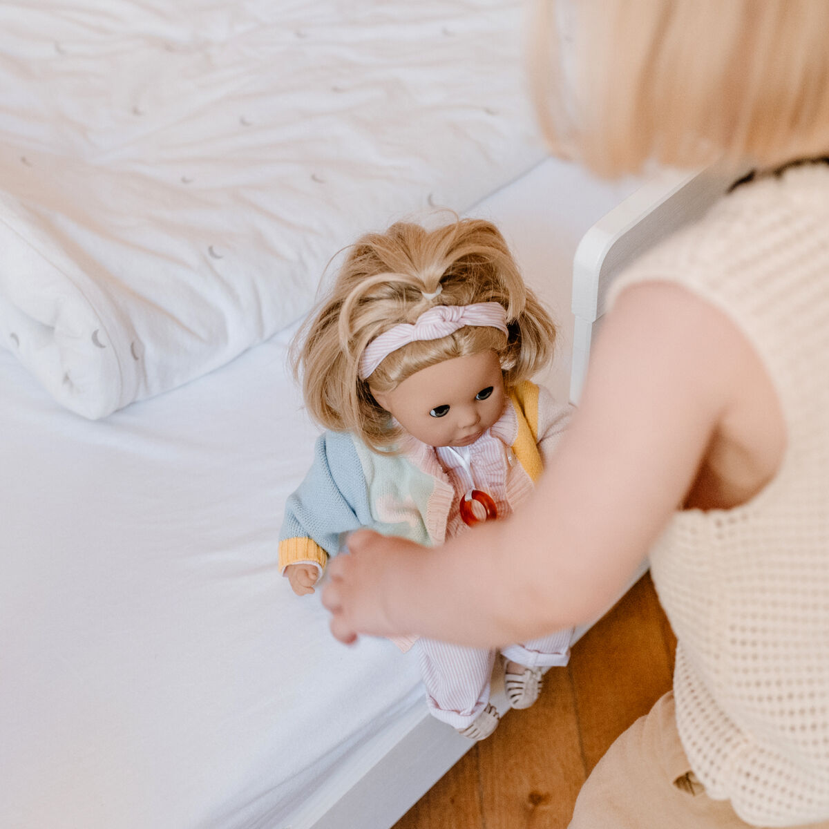 Ein kleines Kind spielt mit einer bunt gekleideten Badepuppe auf einem Bett in einem hellen Kinderzimmer. Die Puppe trägt eine Jacke in Pastellfarben und ein Stirnband, während das Kind ihr interessiert begegnet.