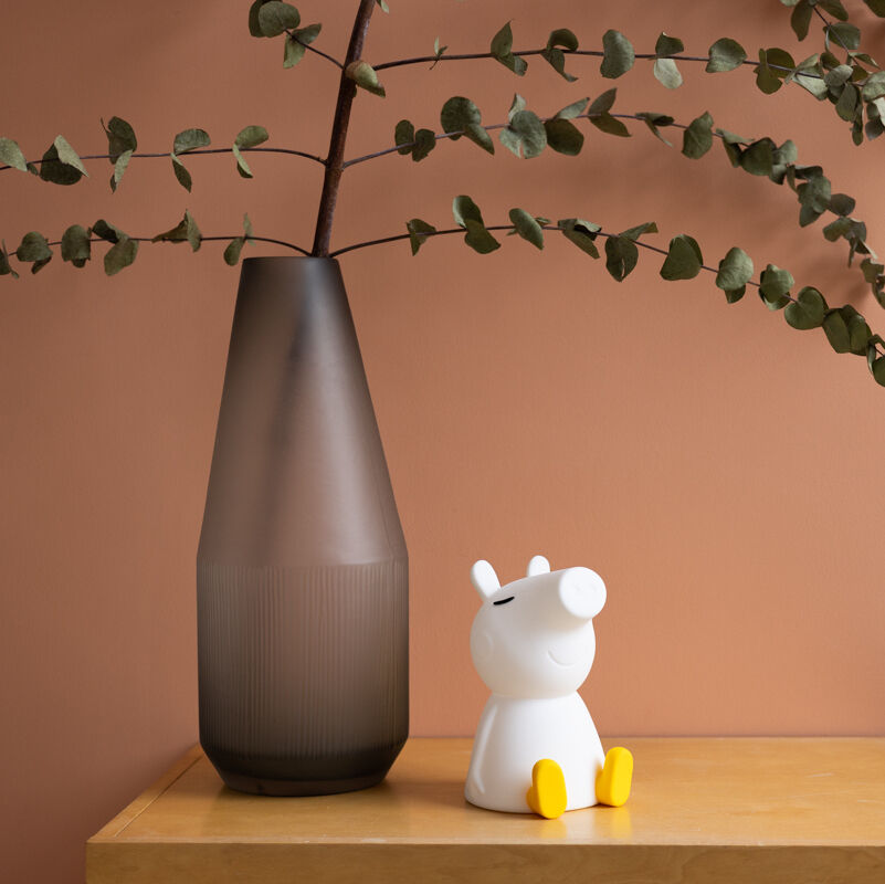 Ein Nachtlicht in Form der Comicfigur Peppa Pig steht auf einem Nachttisch neben einer hohen, braunen Vase mit grünen Zweigen. Der Hintergrund ist eine orangefarbene Wand.