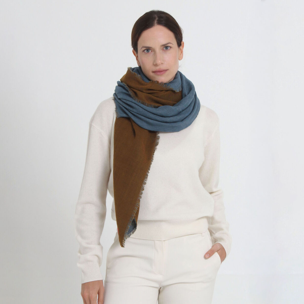 Frau mit Wollschal »Carla« von Shalls Barcelona, bestehend aus 100% Schurwolle, in den Farben Blau und Braun, trägt einen cremefarbenen Pullover und eine helle Hose. Der Schal ist groß, weich, locker um den Hals gelegt und wirkt sehr hochwertig.
