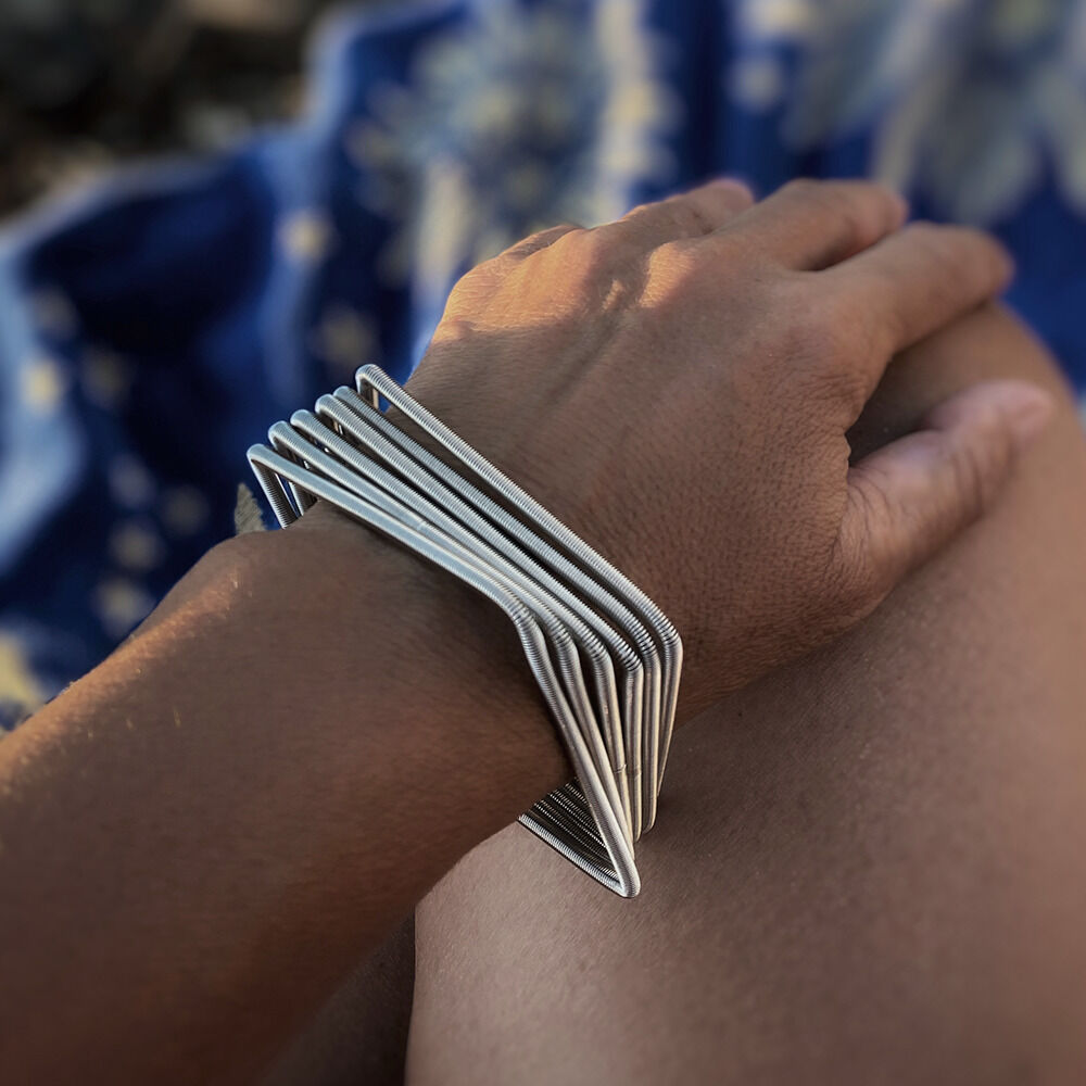Nahaufnahme eines Armbands namens »Square«, das aus verspielten metallischen Quadraten besteht und am Handgelenk einer Person getragen wird. Der Hintergrund zeigt unscharfe blaue und gelbe Muster.
