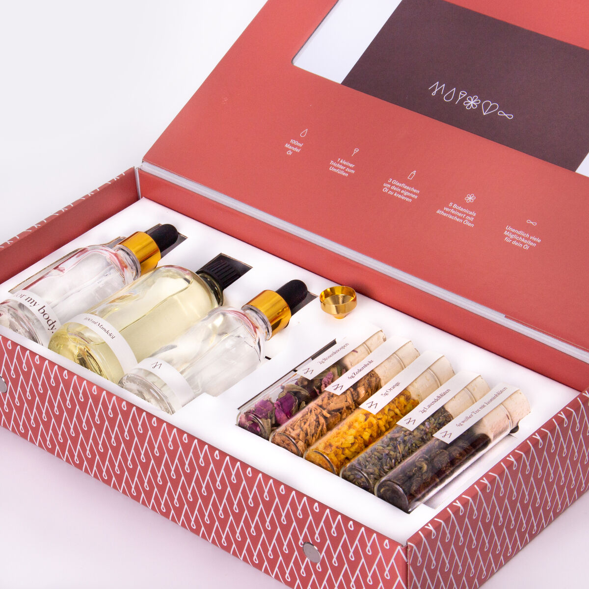 Naturkosmetik-Set MUY in einer eleganten roten Box mit weißen und goldenen Akzenten. Enthält eine Auswahl an Hautpflegeprodukten wie essentielle Öle, Kräuter und weitere Pflegeprodukte in klaren Glasflaschen und Röhrchen. Die Verpackung ist hochwertig und ansprechend gestaltet.