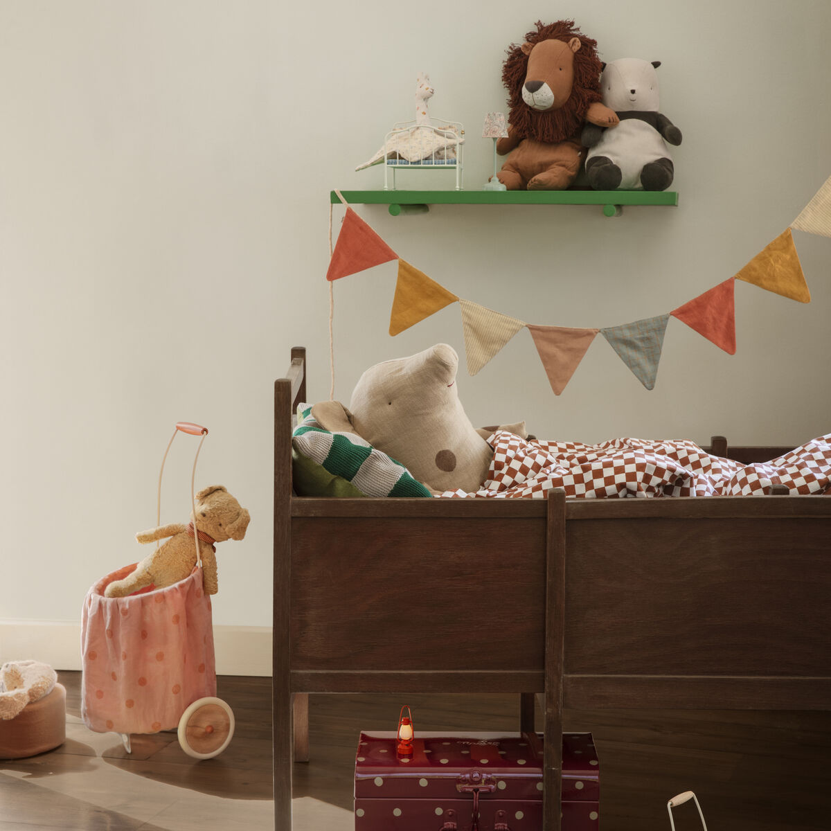 Ein hübsch dekoriertes Babyzimmer mit einem hölzernen Kinderbett und verschiedenen Spielzeugen. Oben auf einem grünen Regal sitzen ein Panda- und ein Löwen-Kuscheltier. Die Wand ist mit einer bunten Wimpelkette geschmückt. Auf dem Boden befinden sich ein Spielzeugauto und ein Tragekorb.