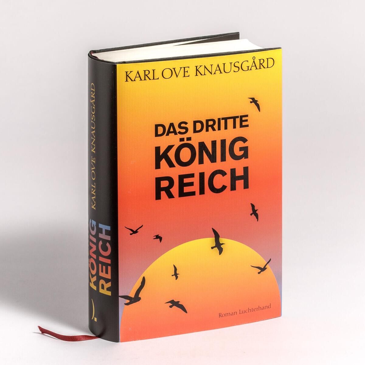 Buchcover von 'Das dritte Königreich' von Karl Ove Knausgård, in Rot- und Schwarztönen mit gelbem Text und stilisierten Vogelsilhouetten auf einem Sonnenuntergangshintergrund. Das Buch hat ein rotes Lesezeichenband aus Stoff.