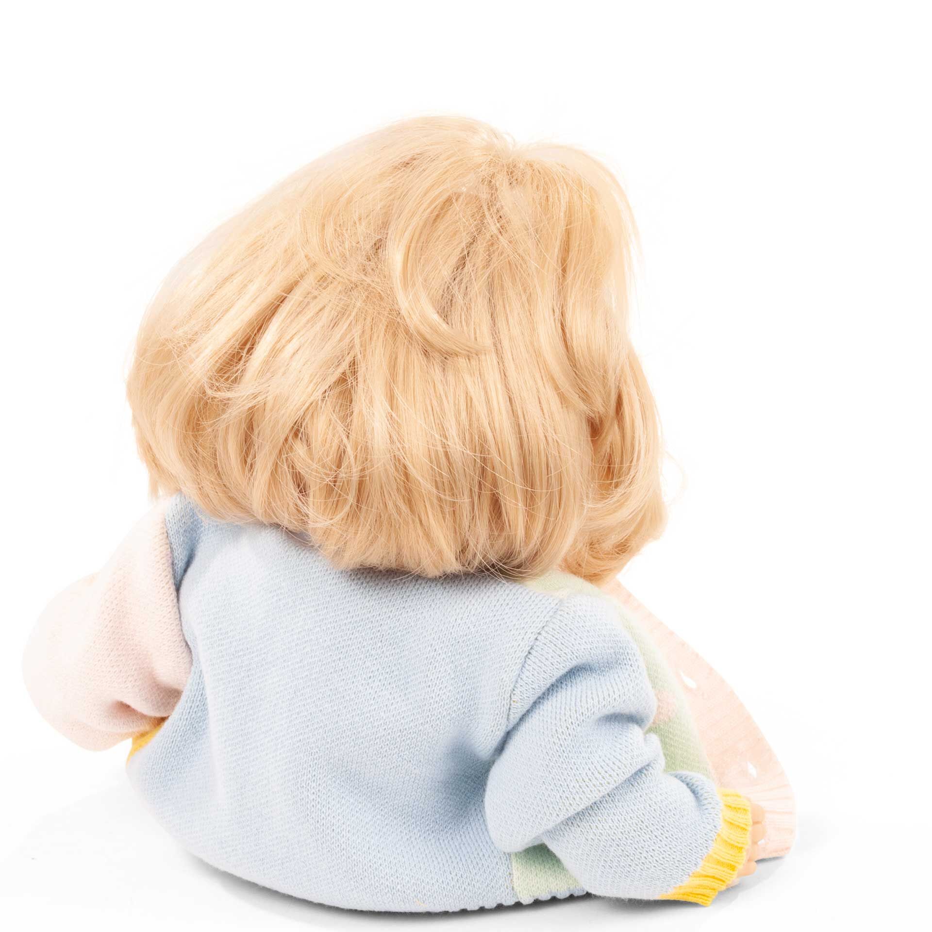 Die Badepuppe »Cosy Aquini Farbspiel« ist von hinten zu sehen. Sie hat schulterlanges blondes Haar und trägt einen bunten Strickpullover in Pastellfarben wie Blau, Rosa, Gelb und Hellgrün. Die Puppe sitzt aufrecht vor einem weißen Hintergrund.