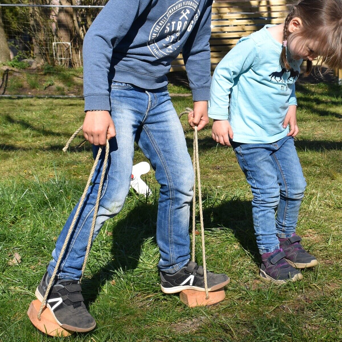 Zwei Kinder spielen draußen im Garten mit Spielhufen. Der Junge trägt einen blauen Pullover und Jeans, das Mädchen trägt ein hellblaues Oberteil und Jeans. Im Hintergrund sind Bäume und ein Haus zu sehen.