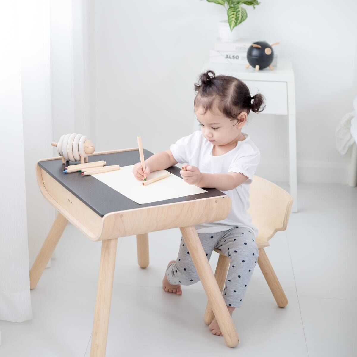 Schwarz-weißer Tisch mit dazu passendem Stuhl aus hellbraunem Holz. Ein kleines Mädchen sitzt am Tisch und malt. Im Hintergrund ist eine weiße Wand zu sehen.