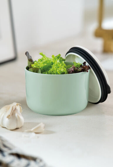 Grüne runde Lunchbox mit Deckel in weiß und schwarz, gefüllt mit frischem grünem Salat, auf einer weißen Oberfläche. Keine anderen Gegenstände bis auf Knoblauchzehen im Bildhintergrund erkennbar.