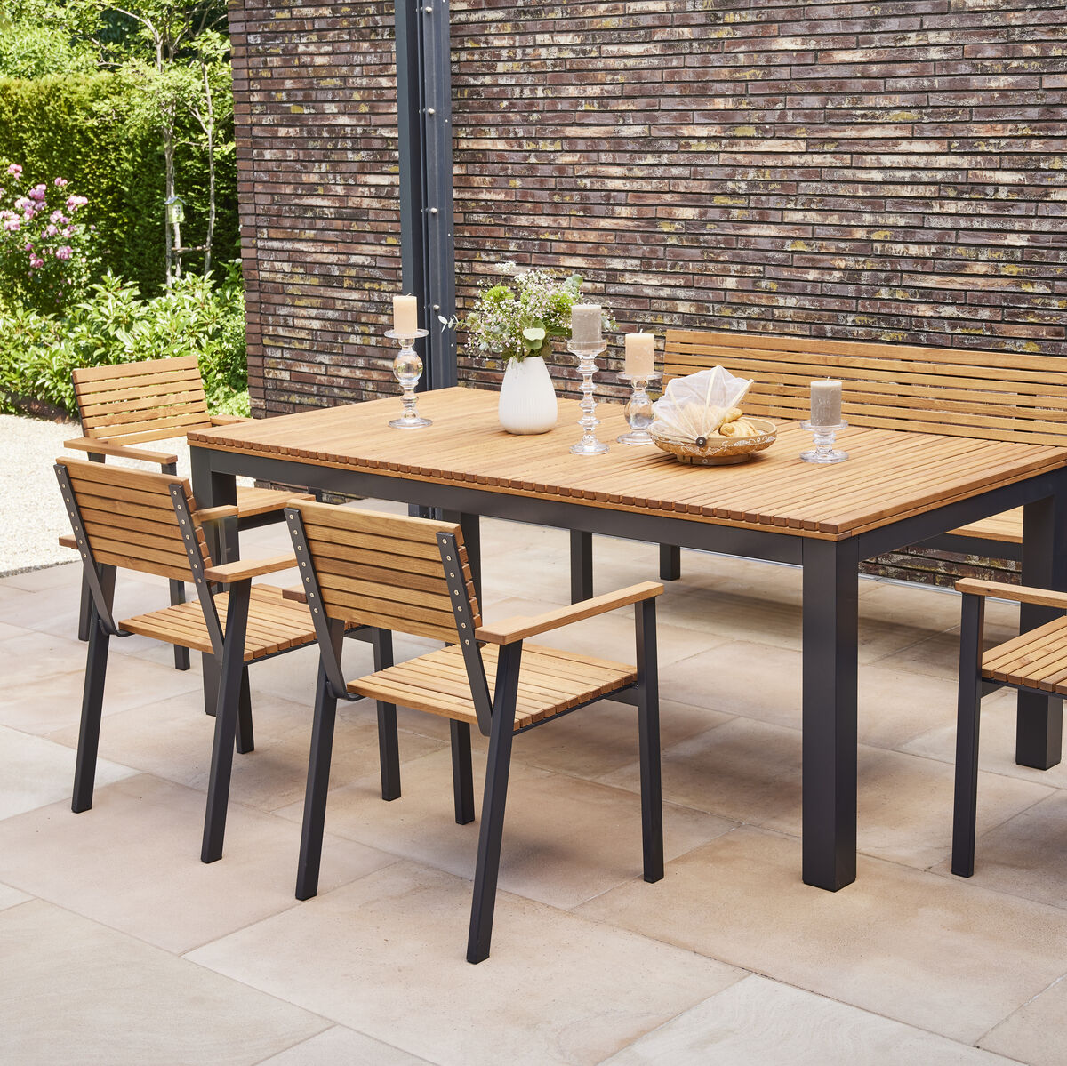 Ein Garten- oder Terrassenset bestehend aus einem rechteckigen Tisch und sechs Stapelsesseln in Holzoptik mit schwarzem Metallrahmen. Das Set befindet sich in einem gepflasterten Außenbereich neben einer Ziegelmauer und umgeben von Pflanzen.