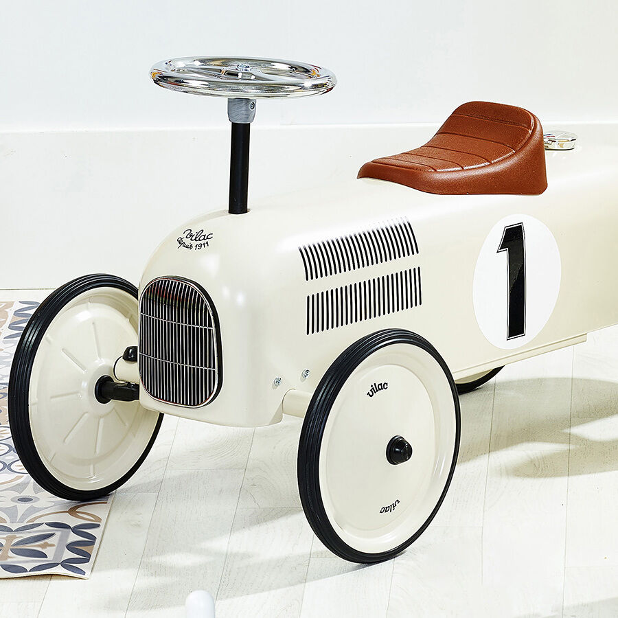 Ein weißes Vintage-Rutschauto aus Metall mit braunem Sitz und der Nummer 1 an der Seite. Es steht auf einem hellen Holzboden vor einer weißen Wand. Das Auto hat schwarze Räder und ein silbernes Lenkrad.