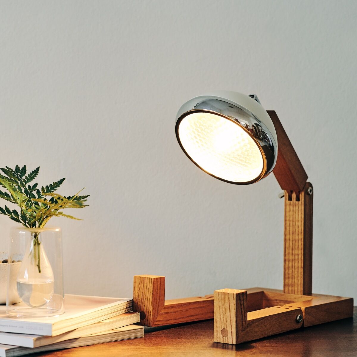 Eine stilvolle Tischlampe im Retro-Design besteht aus Holz und Metall. Sie steht auf einem Holzsockel neben einem Stapel Bücher und einer Vase mit grünen Zweigen.