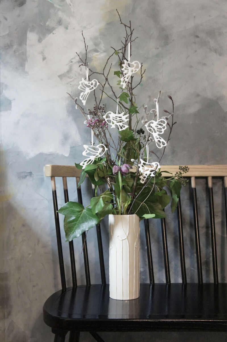 Sechs aus Porzellan gefertigte Vogelornamente hängen an Zweigen in einer Vase, die auf einer schwarzen Holzbank steht. Die Vögel sind kunstvoll und minimalistisch gestaltet, vor einem Hintergrund aus Grautönen und pastellfarbenen Akzenten positioniert.