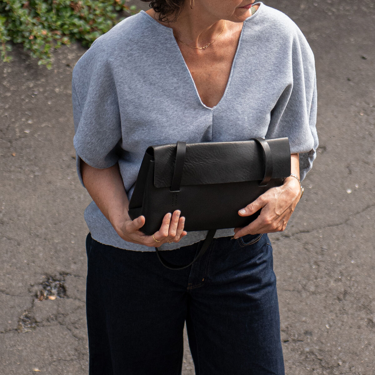 Frau steht auf grauem Asphalt und hält eine schwarze Schultertasche aus hochwertigem Sattlerleder von Harold's Lederwaren GmbH im Arm. Sie trägt einen grauen Pullover mit V-Ausschnitt und eine dunkelblaue Hose. Die Tasche wirkt elegant und hochwertig verarbeitet.