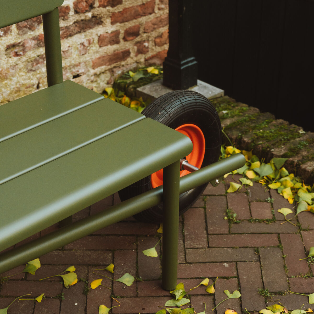 Die Schubkarrenbank »Wheelbench« von Weltevree steht auf braunen Backsteinpflastersteinen, umgeben von herabgefallenem gelbem Laub. Ein Teil der Bank sowie das schwarze Rad mit orangefarbener Felge sind sichtbar. Neben der Bank eine Backsteinmauer und ein schwarzes Holztor.