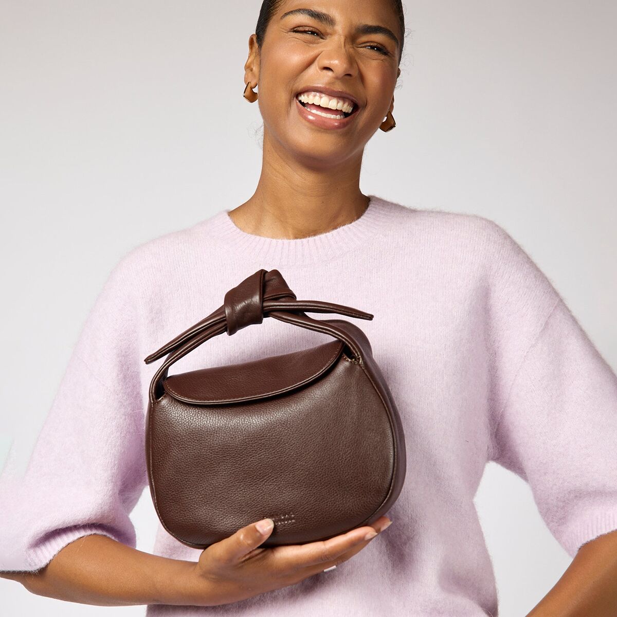 Eine elegante, halbmondförmige Tasche mit geschwungenem Überschlag aus luxuriösem schokoladenbraunem Soft-Narbenleder wird von einer Person vor einem blassrosa Strickpullover präsentiert. Die Tasche wirkt hochwertig verarbeitet, die Oberfläche ist fein genarbt und leicht glänzend.