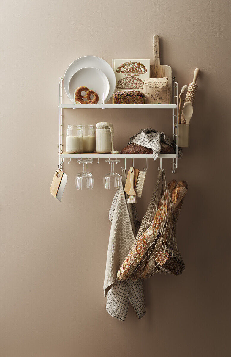 Ein weißes String Küchenregal an einer beige gestrichenen Wand, dekoriert mit Milchflaschen, Brot, Küchenutensilien, Glass Gläsern und einer Netztasche mit Baguette.