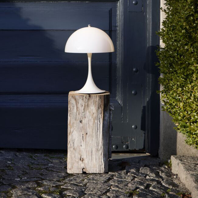 Ein Bild zeigt die tragbare Leuchte 'Panthella 250' in Weiß aus Acryl, die draußen auf einem hölzernen Stamm steht. Im Hintergrund ist eine dunkelgraue Tür und ein Busch zu sehen.