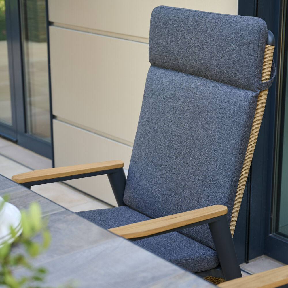 Ein moderner Sessel mit grauer Polsterung und Holzarmlehnen, geeignet für den Einsatz auf Terrassen und in Esszimmern. Der Sessel hat eine hohe Rückenlehne und zieht sich stilvoll in einer Outdoor-Umgebung mit Glasfenstern und Holzdetails an.